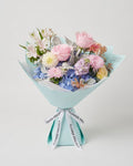A pastel flower bouquet wrapped in light blue paper with ribbon printed with 'FLOWER PROJECT', featuring pink roses, blue hydrangeas, and purple stock flowers on a light gray background.