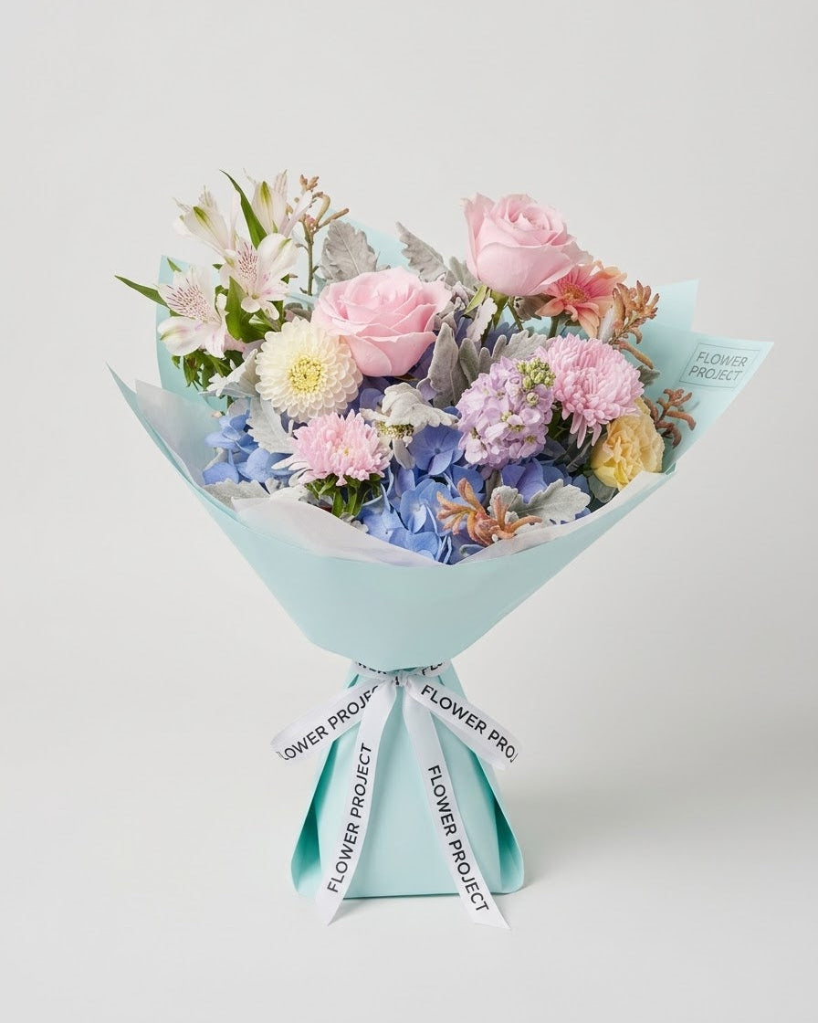 A pastel flower bouquet wrapped in light blue paper with ribbon printed with 'FLOWER PROJECT', featuring pink roses, blue hydrangeas, and purple stock flowers on a light gray background.