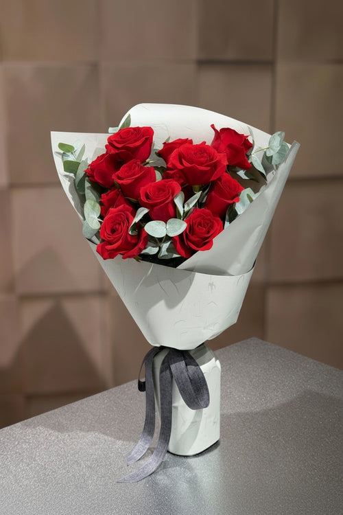 A bouquet of 12 red roses wrapped in white paper with green eucalyptus leaves and tied with a gray ribbon.