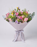 A beautifully arranged floral bouquet featuring pink roses, carnations, purple flowers, and green foliage, wrapped in light grey paper with a matching ribbon tied in a bow, set against a solid light gray background