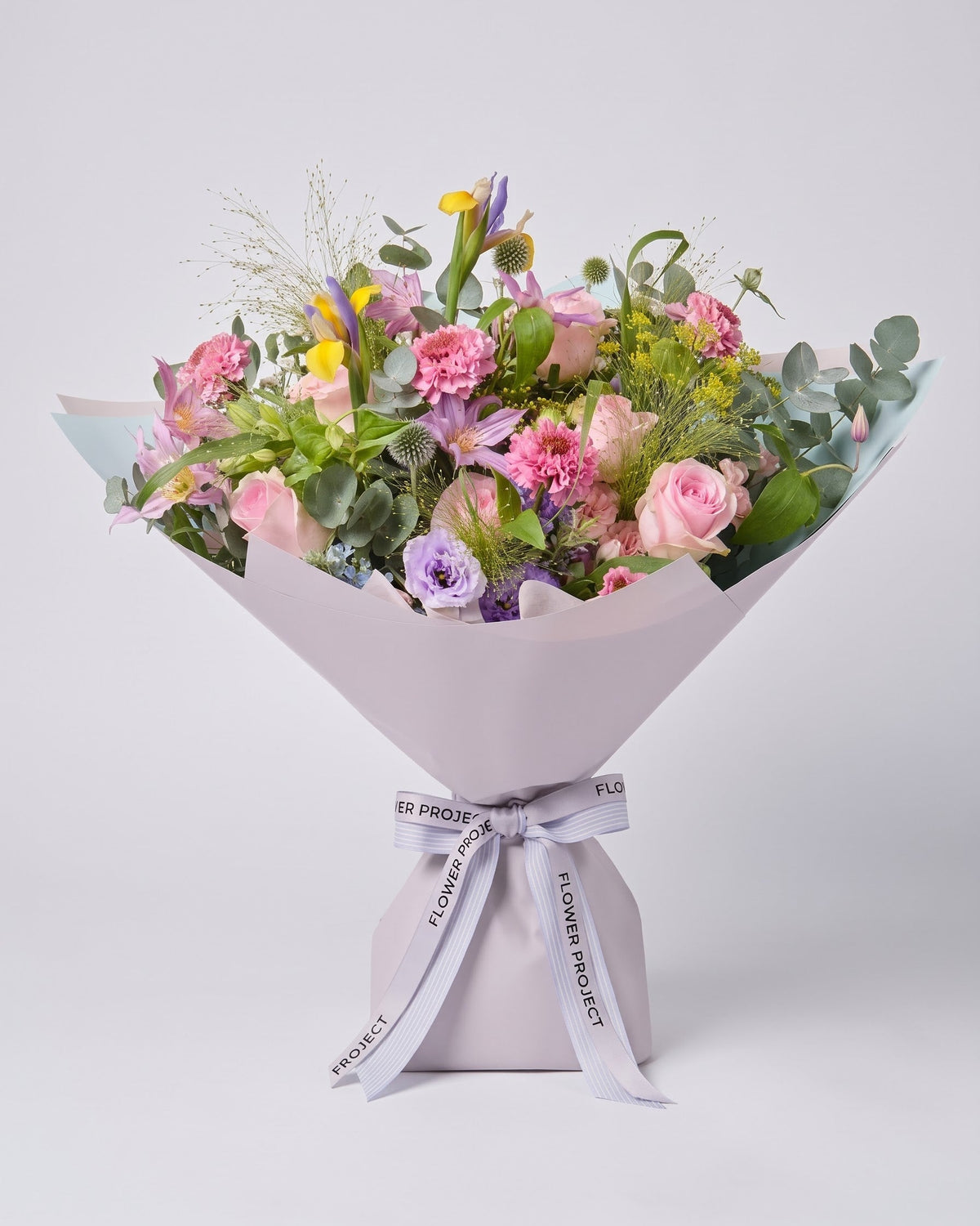 A beautifully arranged floral bouquet featuring pink roses, carnations, purple flowers, and green foliage, wrapped in light grey paper with a matching ribbon tied in a bow, set against a solid light gray background